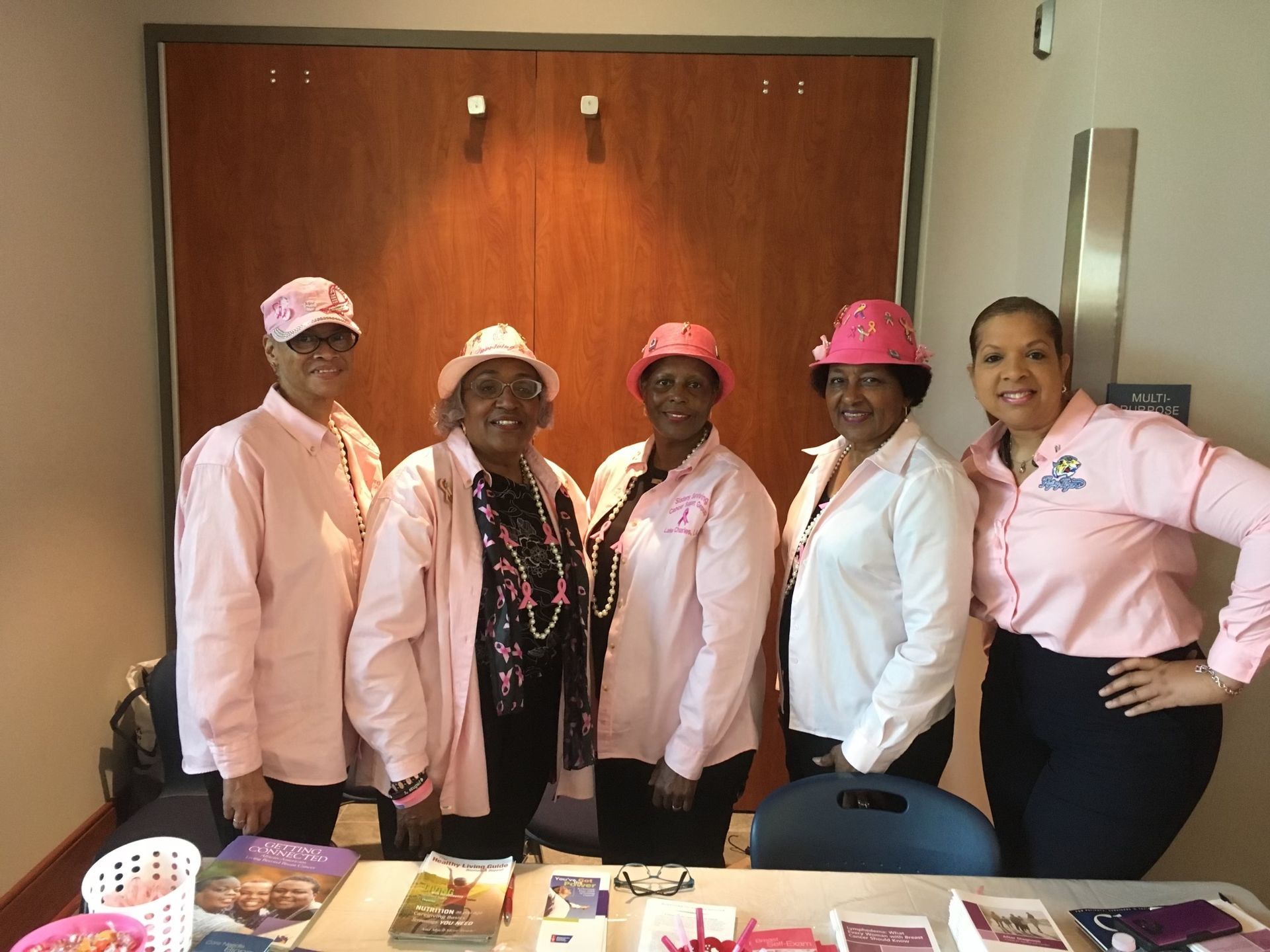 A group of women wearing pink shirts and pink hats are standing around a table.
