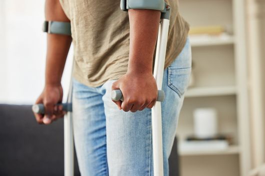 Person using crutches, indoors. Light blue jeans, tan shirt. Holding crutch handles, with focus on arms and hands.