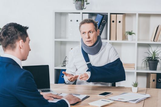 Man in arm sling and neck brace speaking to a person at a desk in an office setting.