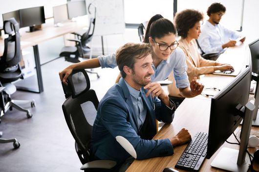 People working at computers in an office setting. Woman pointing at screen, smiling.