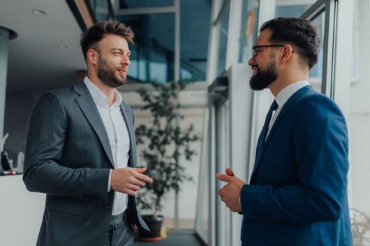 Two men in suits conversing near a window in a modern office.