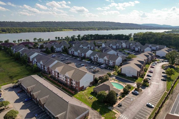 Aerial view of apartment complex with pool, parked cars, and a lake in the background.