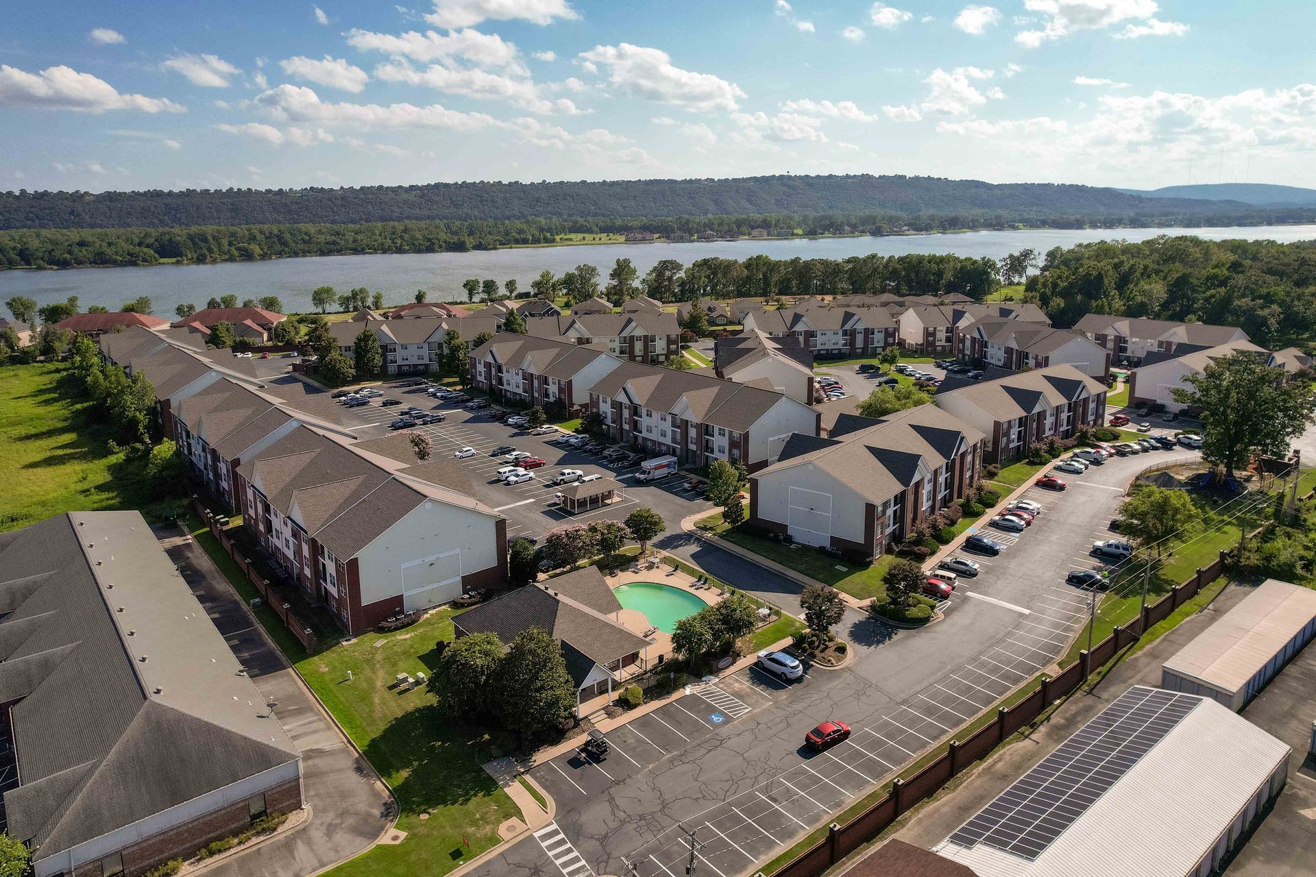 Aerial view of an apartment complex with a pool, parking lot, and lakefront view on a sunny day.