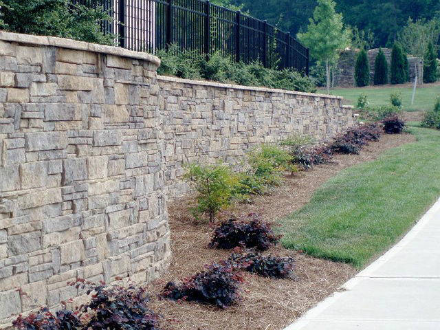 A stone wall along a sidewalk with a fence in the background.