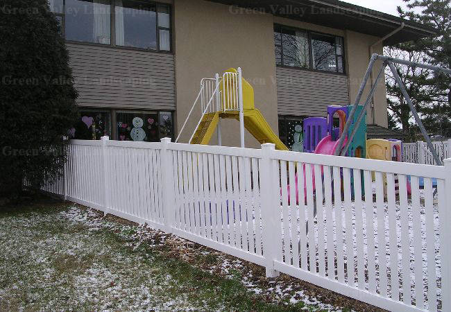 A white fence surrounds a playground in front of a building