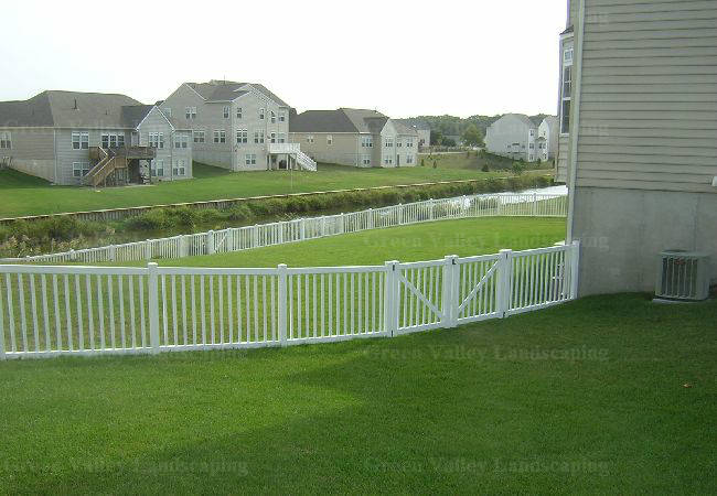 A white fence surrounds a lush green yard with houses in the background