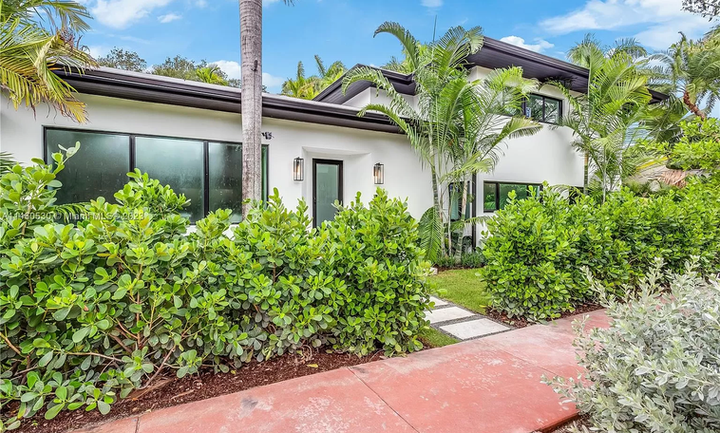 A white house with a red walkway leading to it is surrounded by trees and bushes.