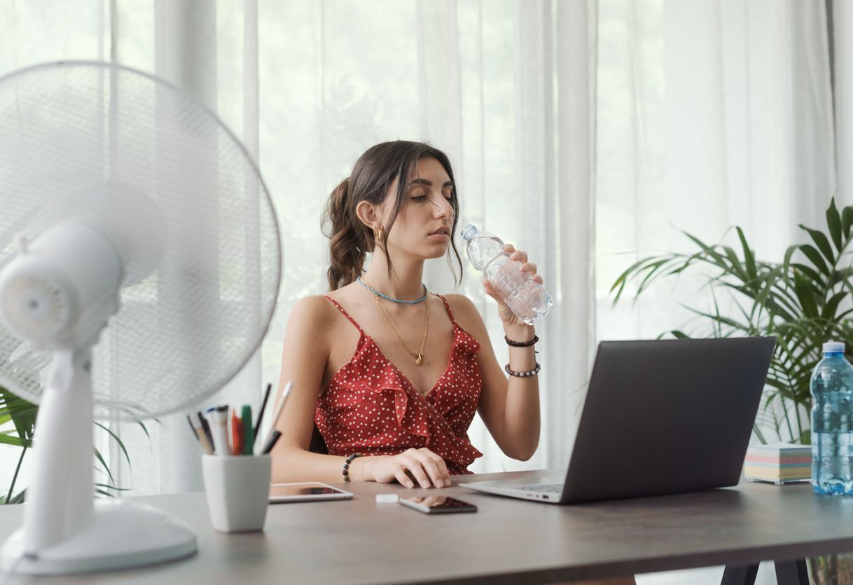 Woman at a desk drinking water, facing a fan, working on a laptop in a bright room.