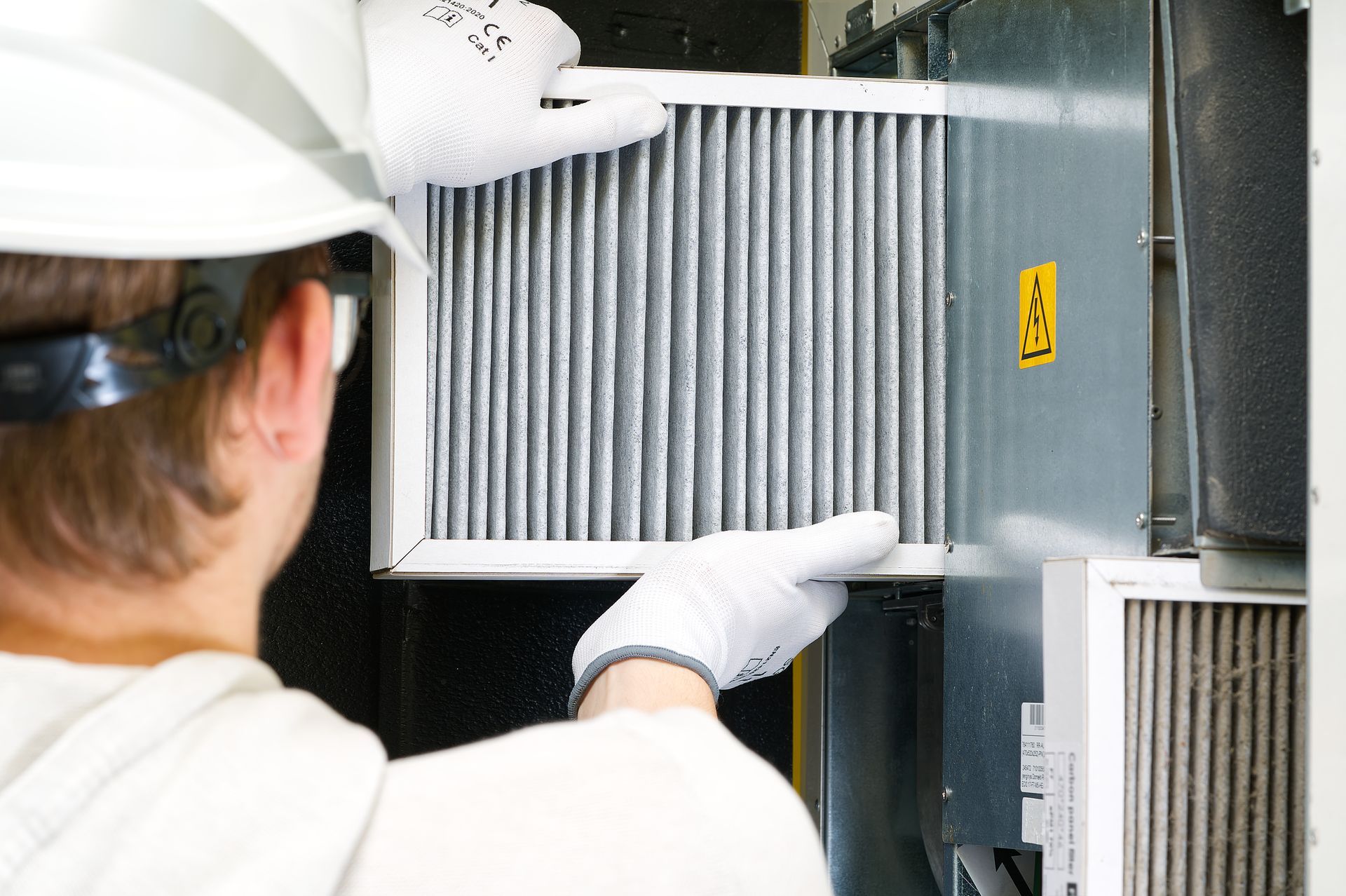 Person in gloves and hard hat replacing an air filter in an HVAC unit.