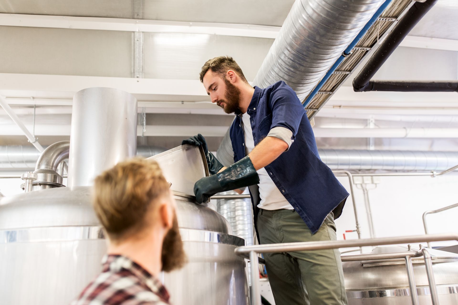 Two men brewing beer, one adding ingredients to a large steel vat, another watching in a brewery.