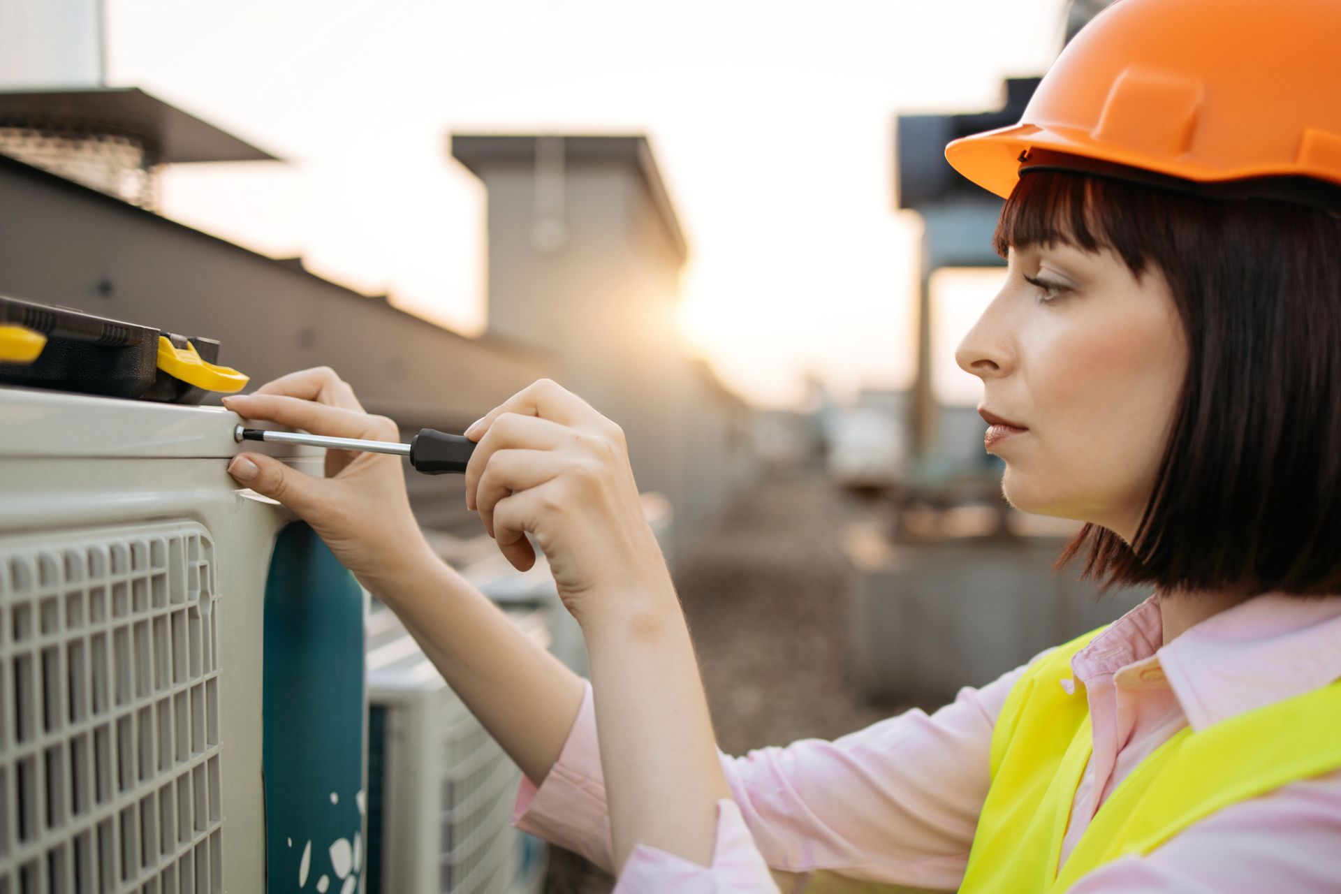 Woman in hard hat and safety vest repairs HVAC unit with a screwdriver outdoors.