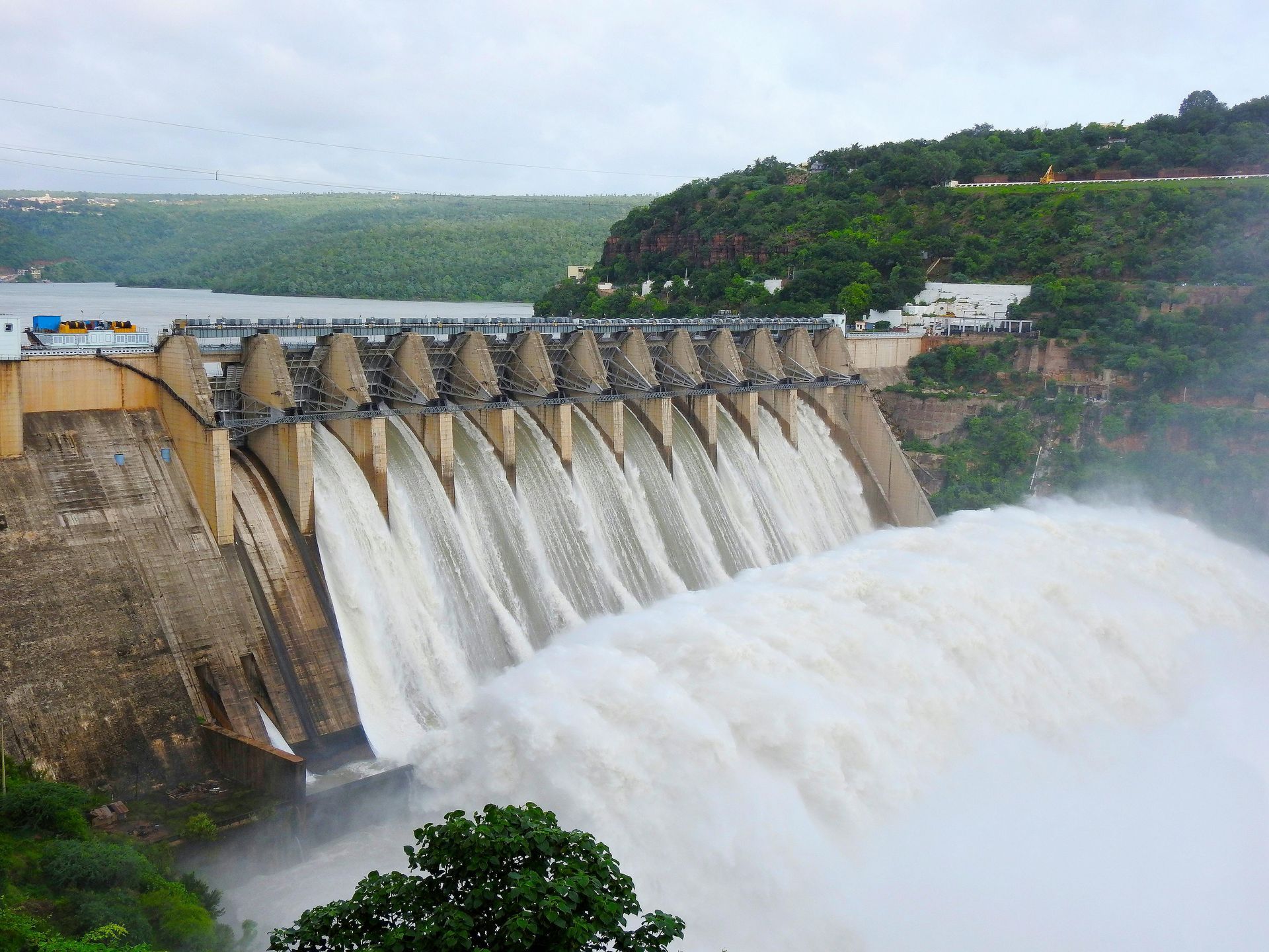 Dam releasing water; brown structure with white water cascading below, forested hills in the background.