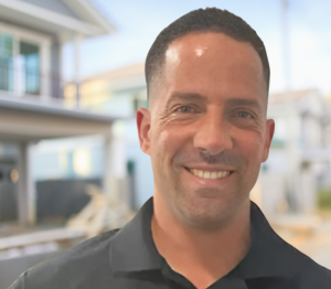 A man in a black shirt is smiling in front of a house.