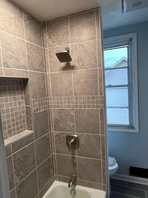 Shower with gray tile, a built-in shelf, and a window next to the toilet.