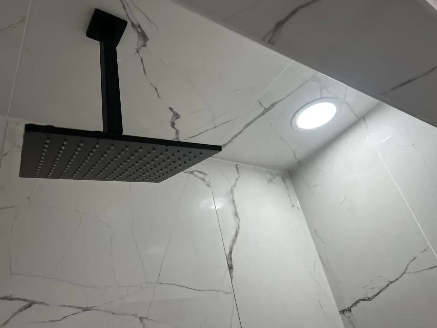 Black square rain showerhead and recessed ceiling light in a white tiled shower.