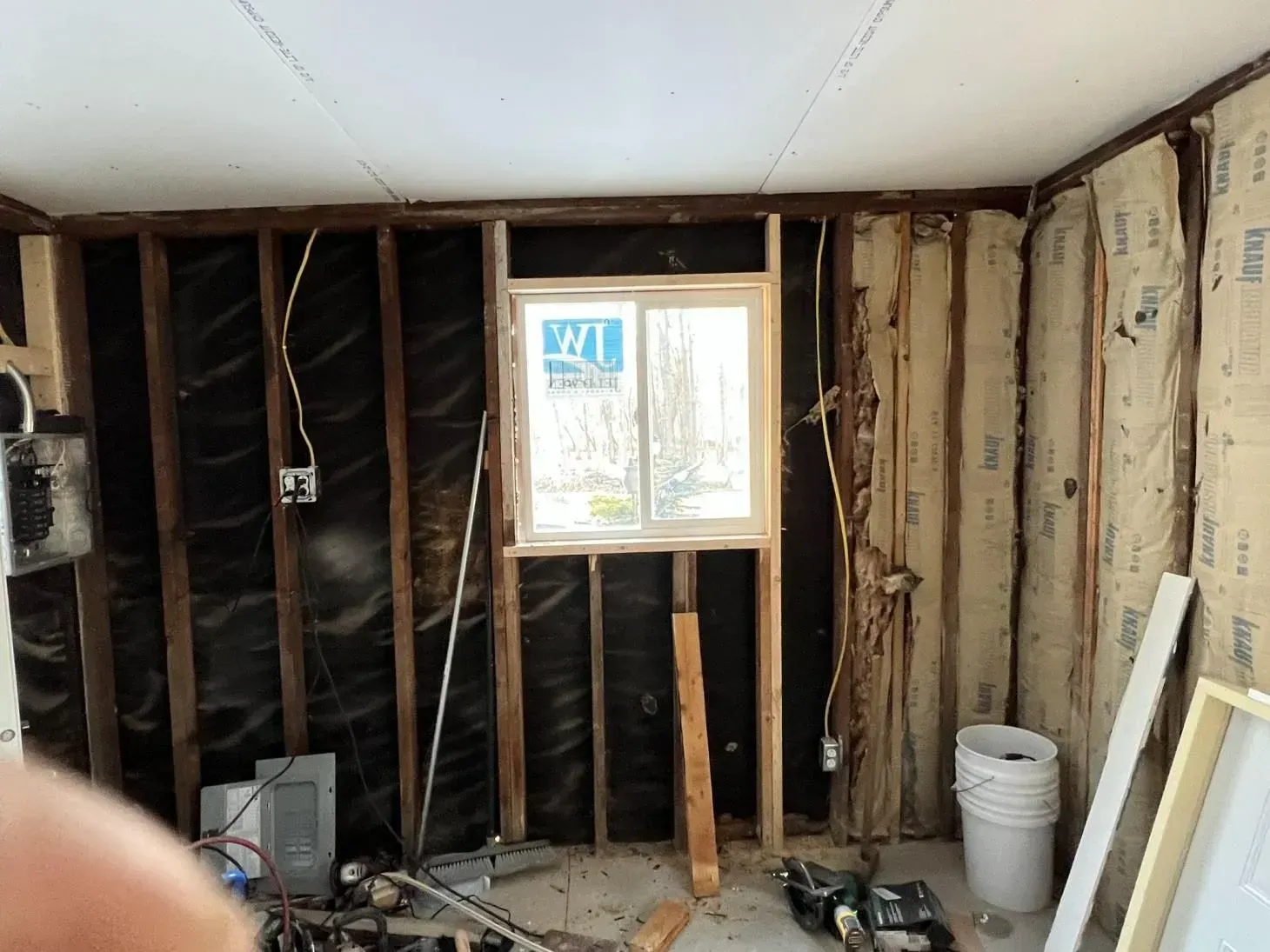 Interior view of a room under renovation. Exposed wooden studs, insulation, a window, and electrical wiring are visible.