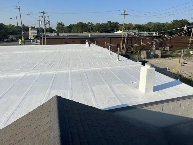 White-coated commercial roof with a chimney, seen from an adjacent rooftop, sunny day.