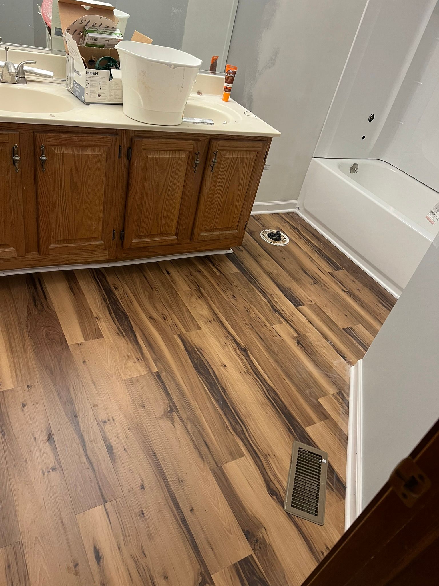 Bathroom with wood-look flooring, wooden vanity, white tub, and a white bucket.
