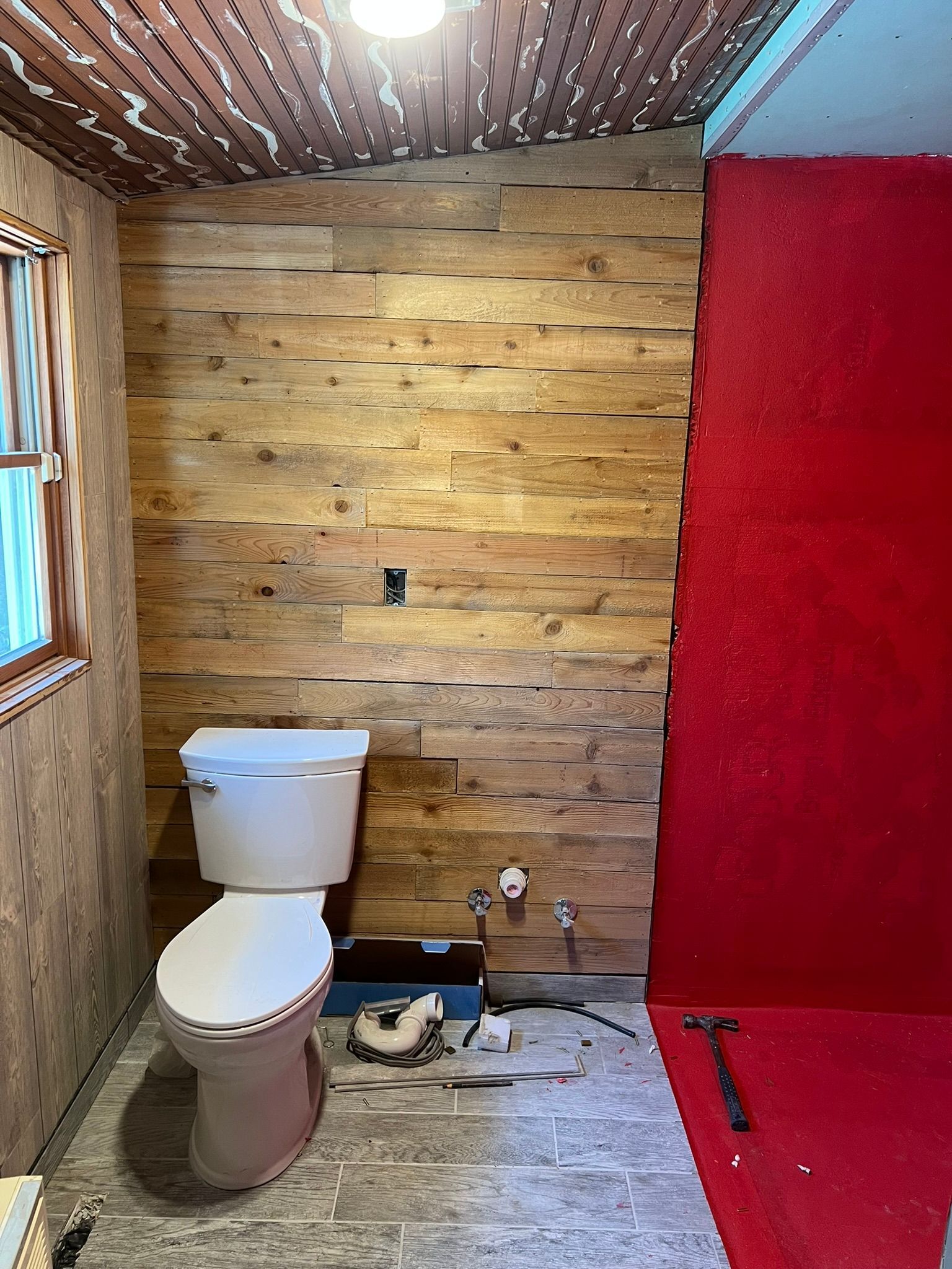 Bathroom renovation with wood-paneled wall, toilet, and red-painted wall. Unfinished flooring, tools present.