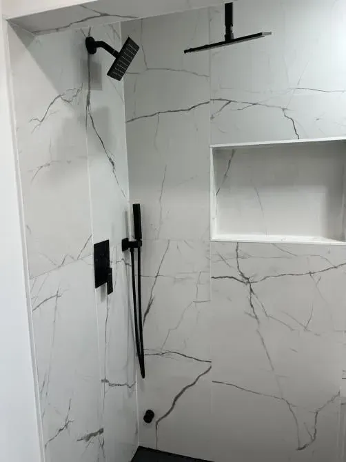 Modern white and gray tiled shower with black fixtures, including shower heads, a handheld wand, and a recessed shelf.