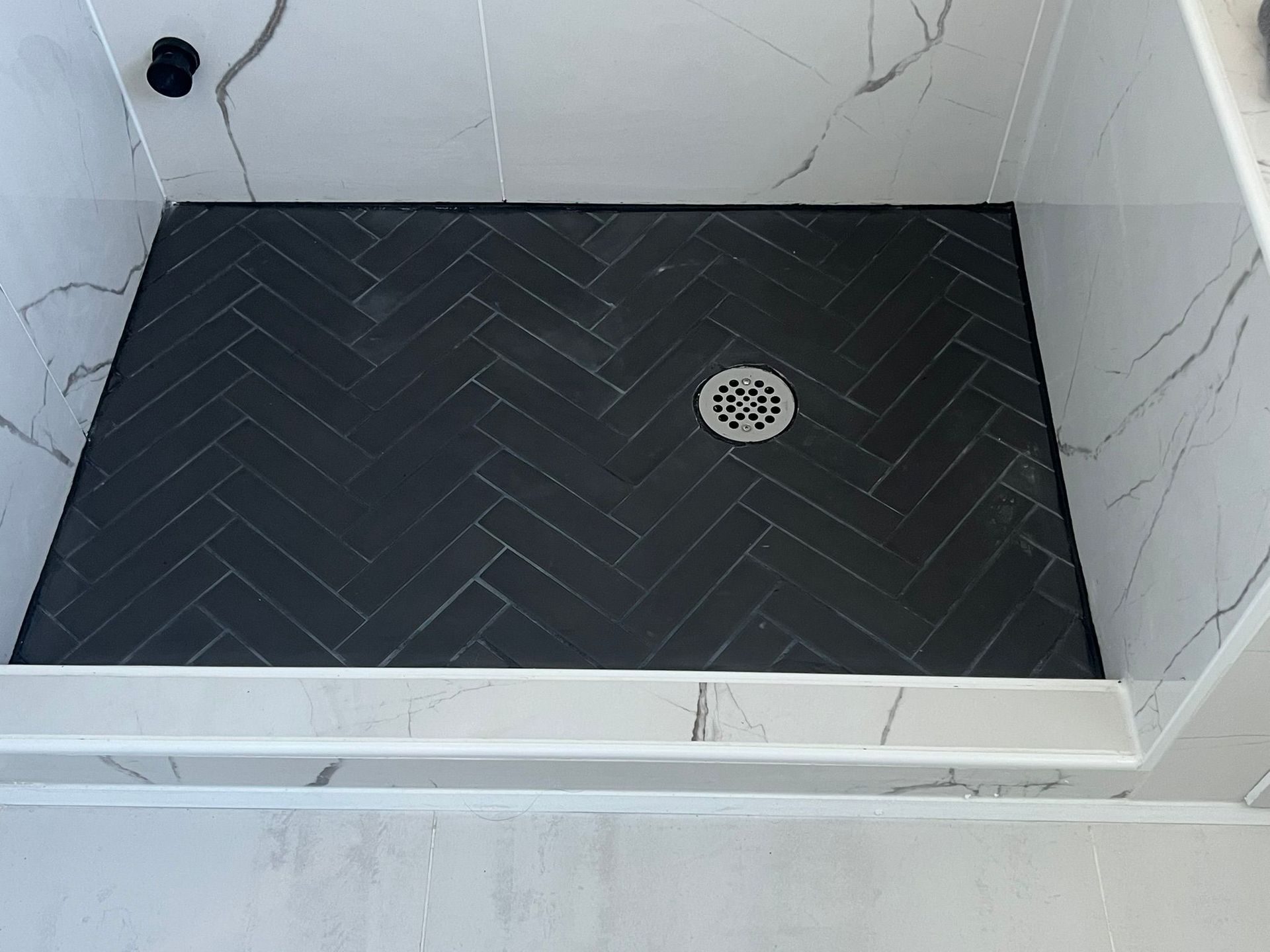 Black herringbone tile shower floor with drain, set in marble-patterned walls.