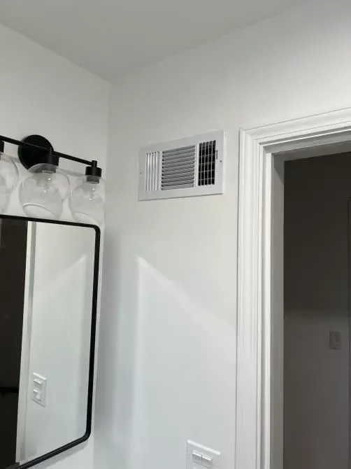 White bathroom wall with air vent, mirror, and open doorway.