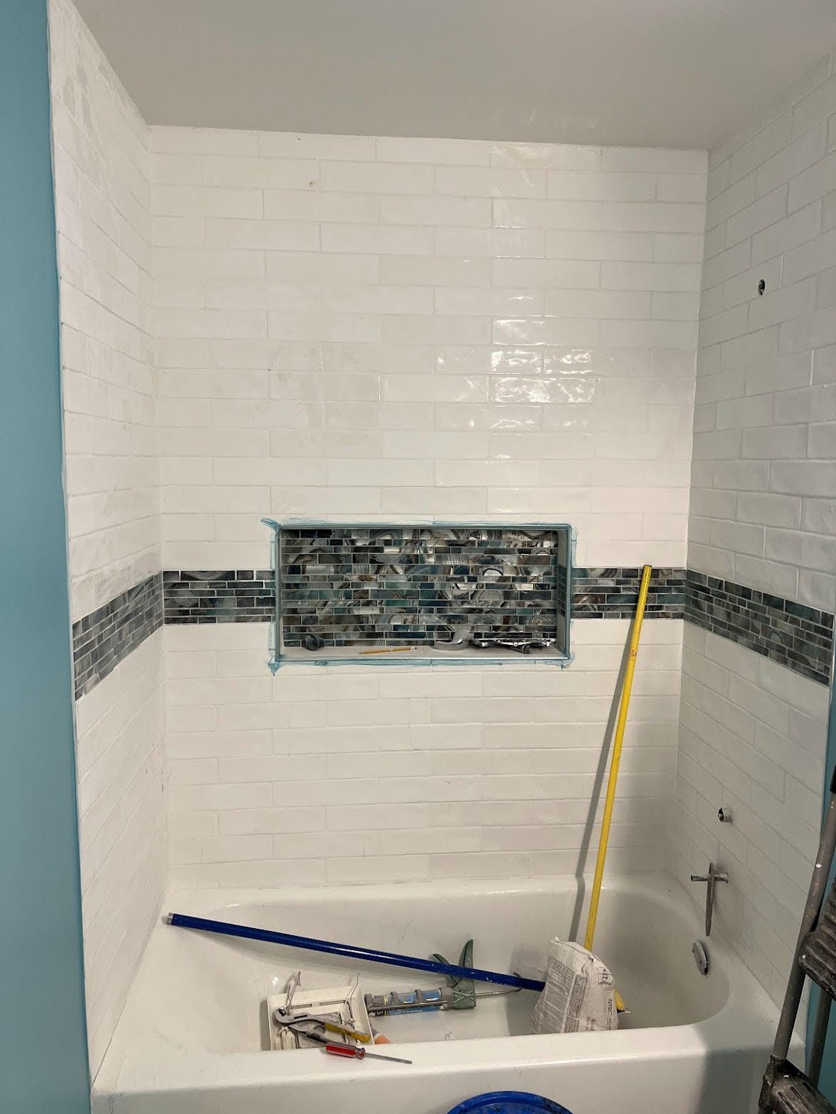 Bathroom remodel: White tiled walls with a dark mosaic band. Bathtub and niche with blue-gray accent tiles.