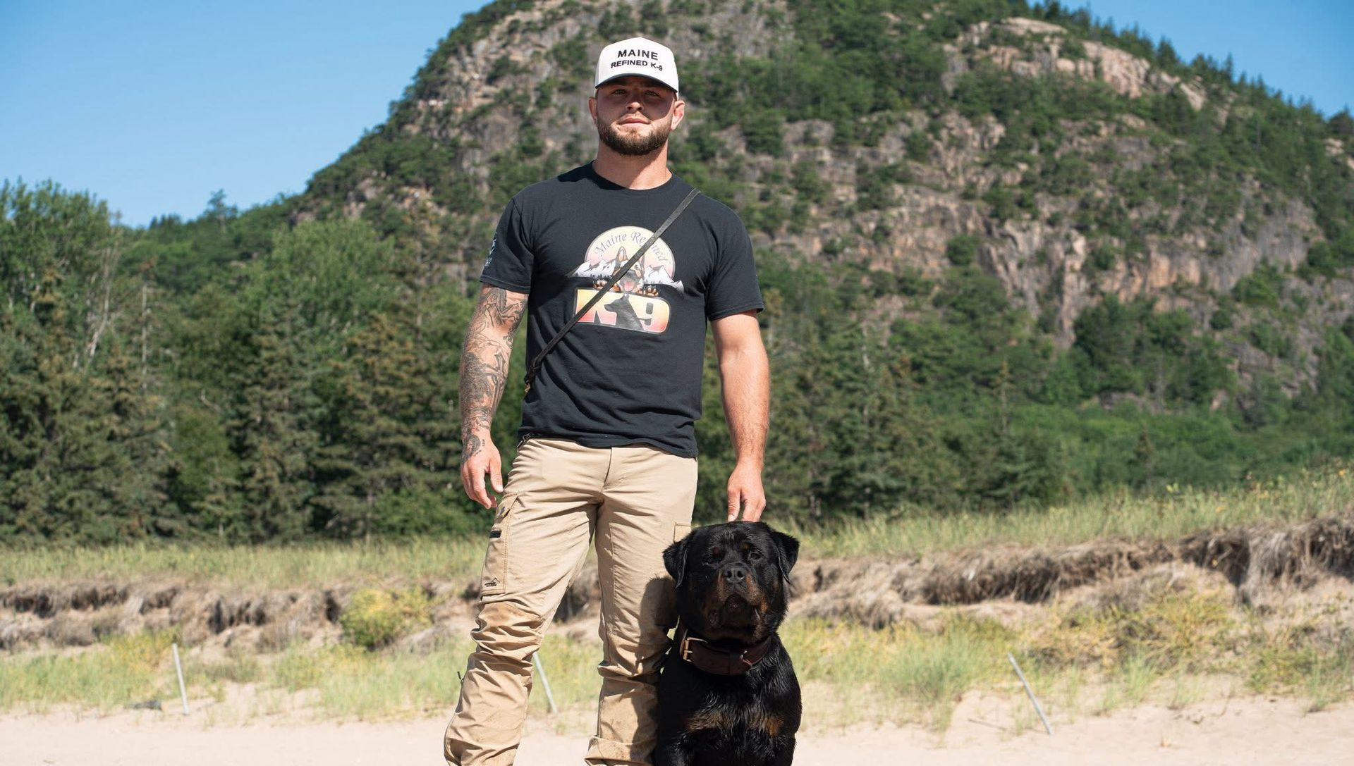 Man in cap and t-shirt with dog on sandy beach, mountain in background.