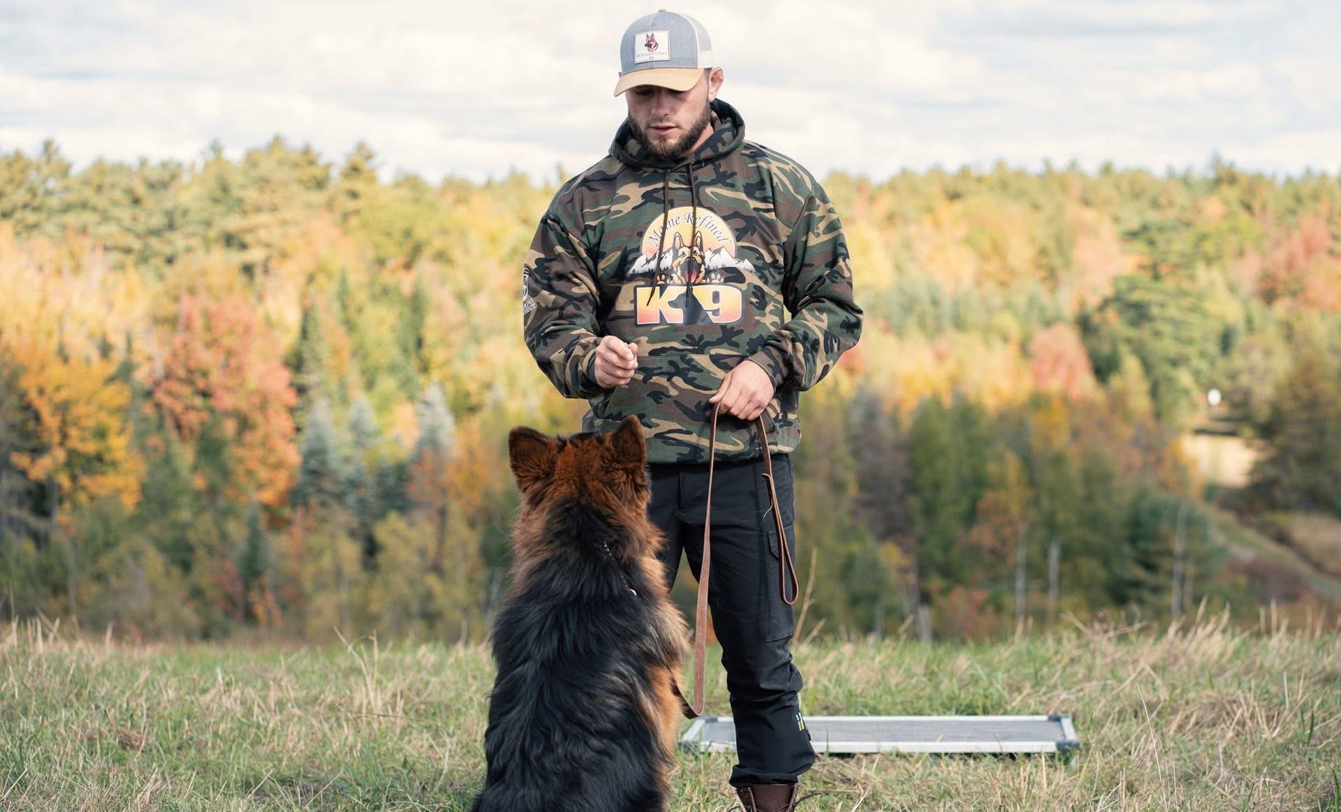 Man in camo hoodie training a German Shepherd dog outdoors.