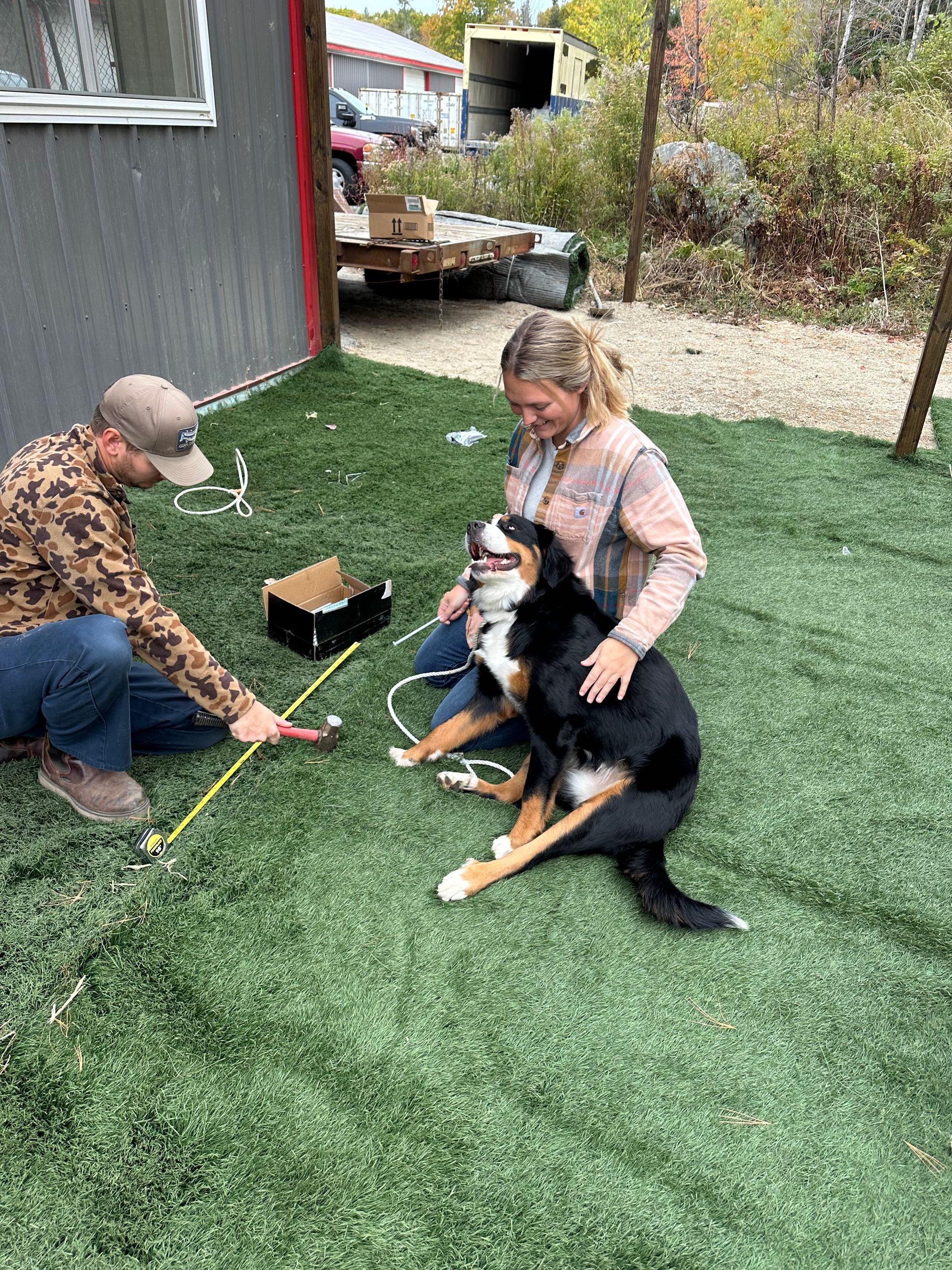 Man plays with dog on turf while woman pets it, outdoors.