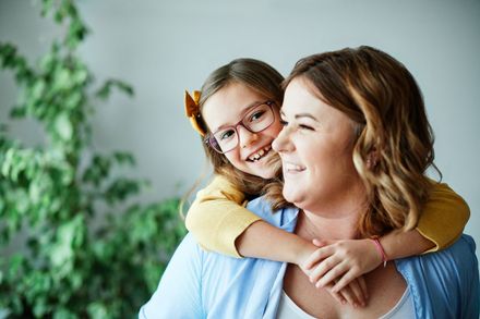 Girl wearing glasses hugs a woman with a smile, indoor setting, plant in background.