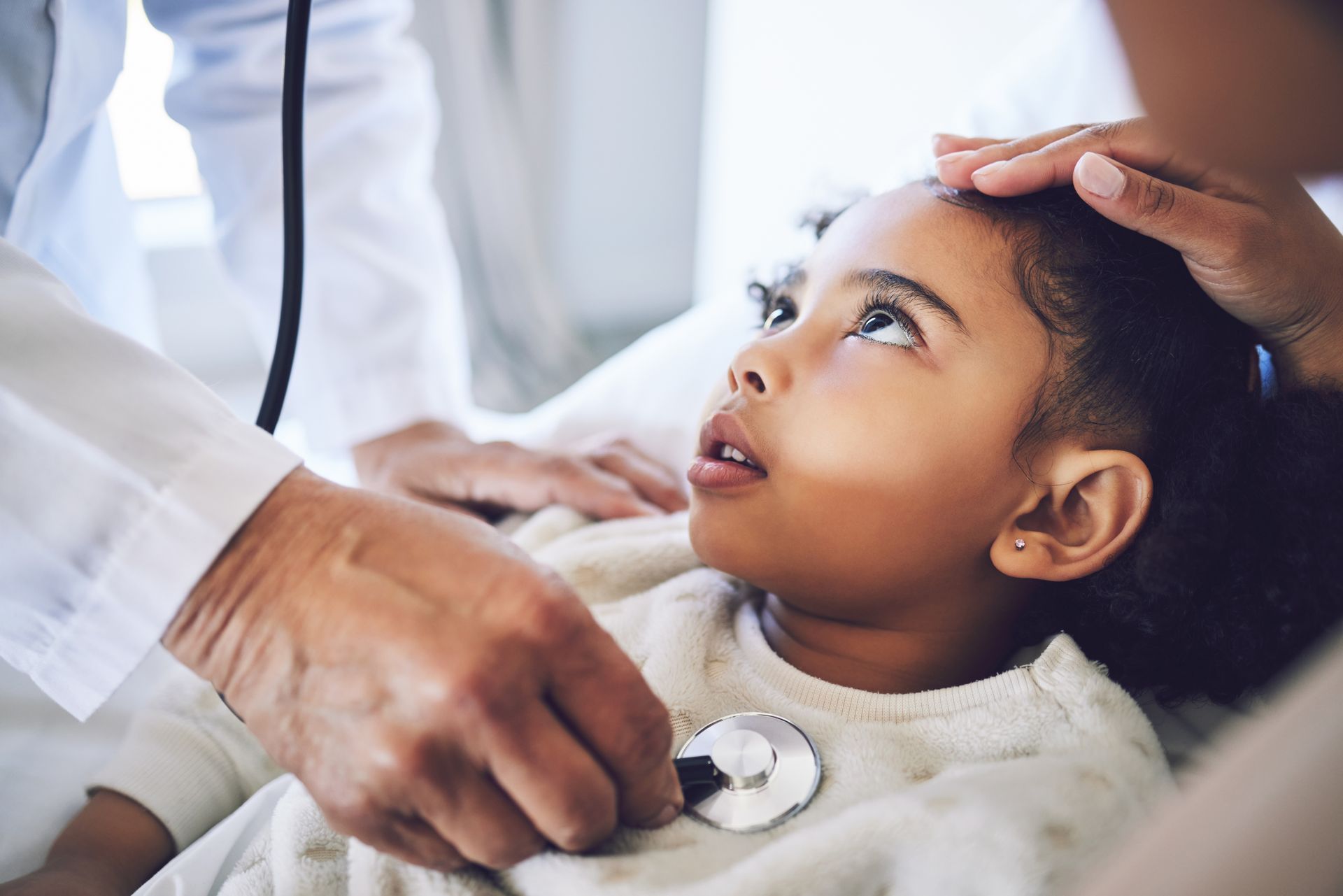 Doctor listening to a child's chest with a stethoscope while a parent touches the child's forehead.