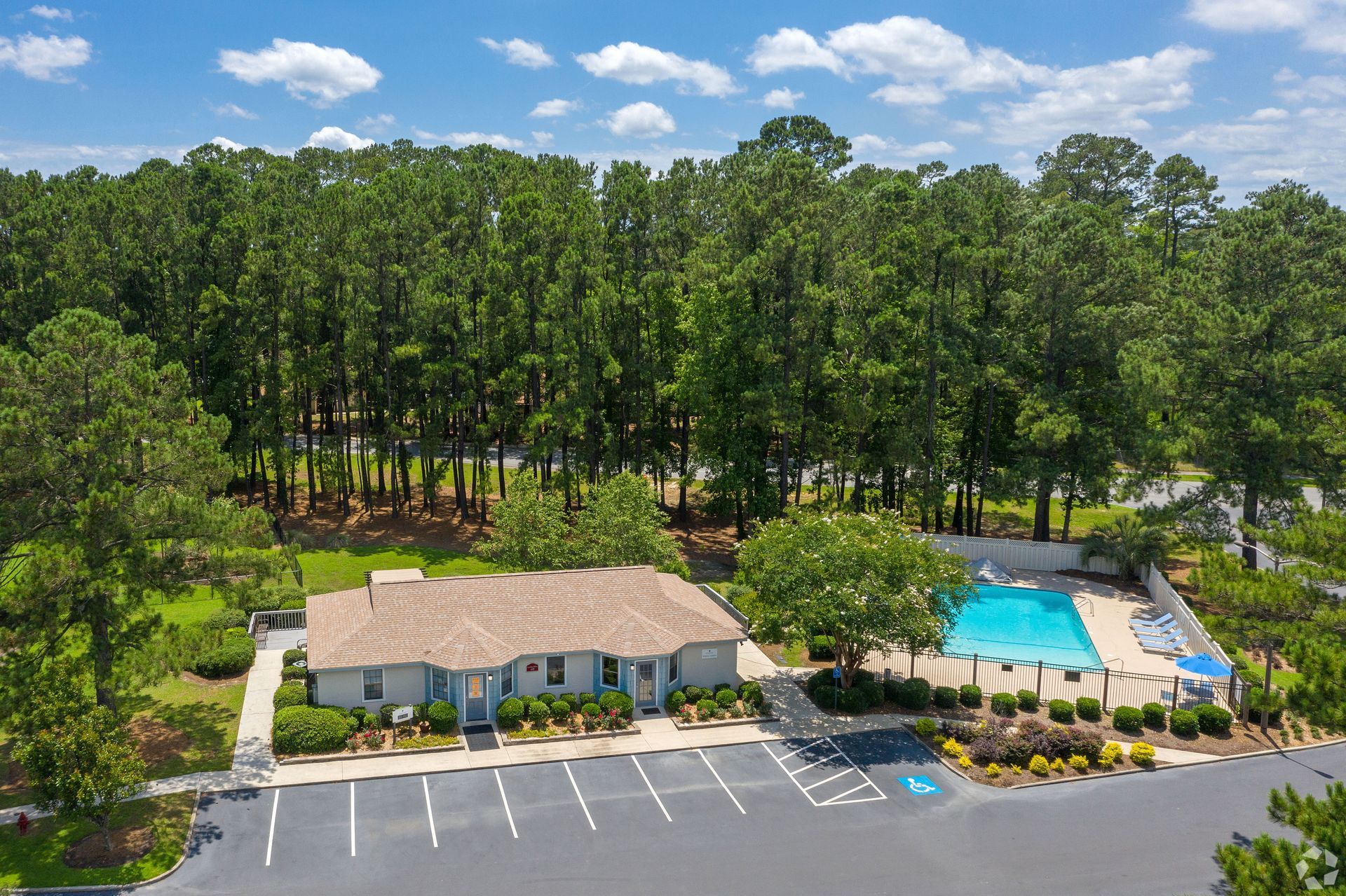 A building with a pool and parking lot in front of a treeline on a sunny day.