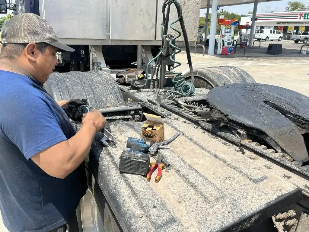 Man working on a semi-truck's fifth wheel. Tools and parts are on the dirty surface near a gas station.