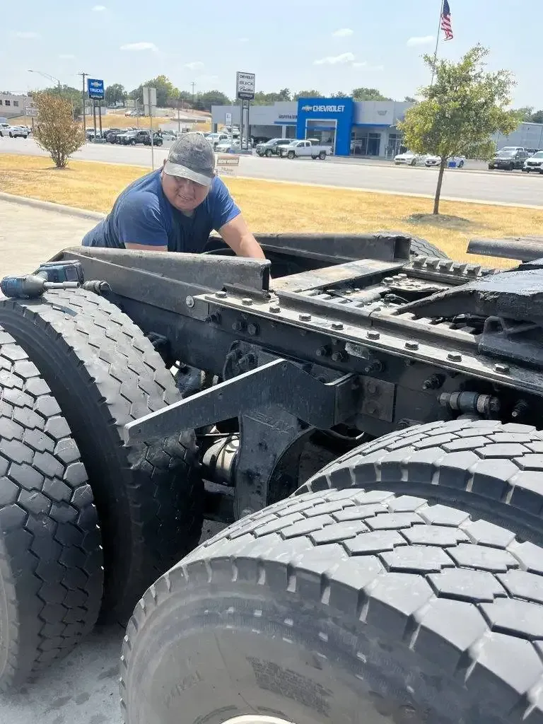 Person inspecting the undercarriage of a semi-truck; outdoors, sunny day.