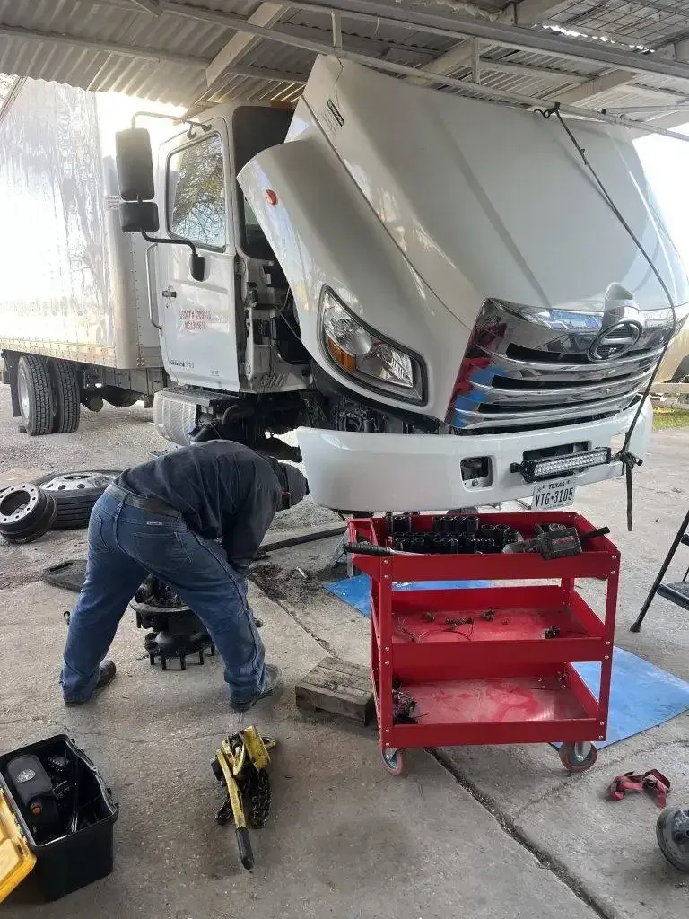 Mechanic working on a white truck with open hood; red toolbox on wheels, tools, indoor setting.