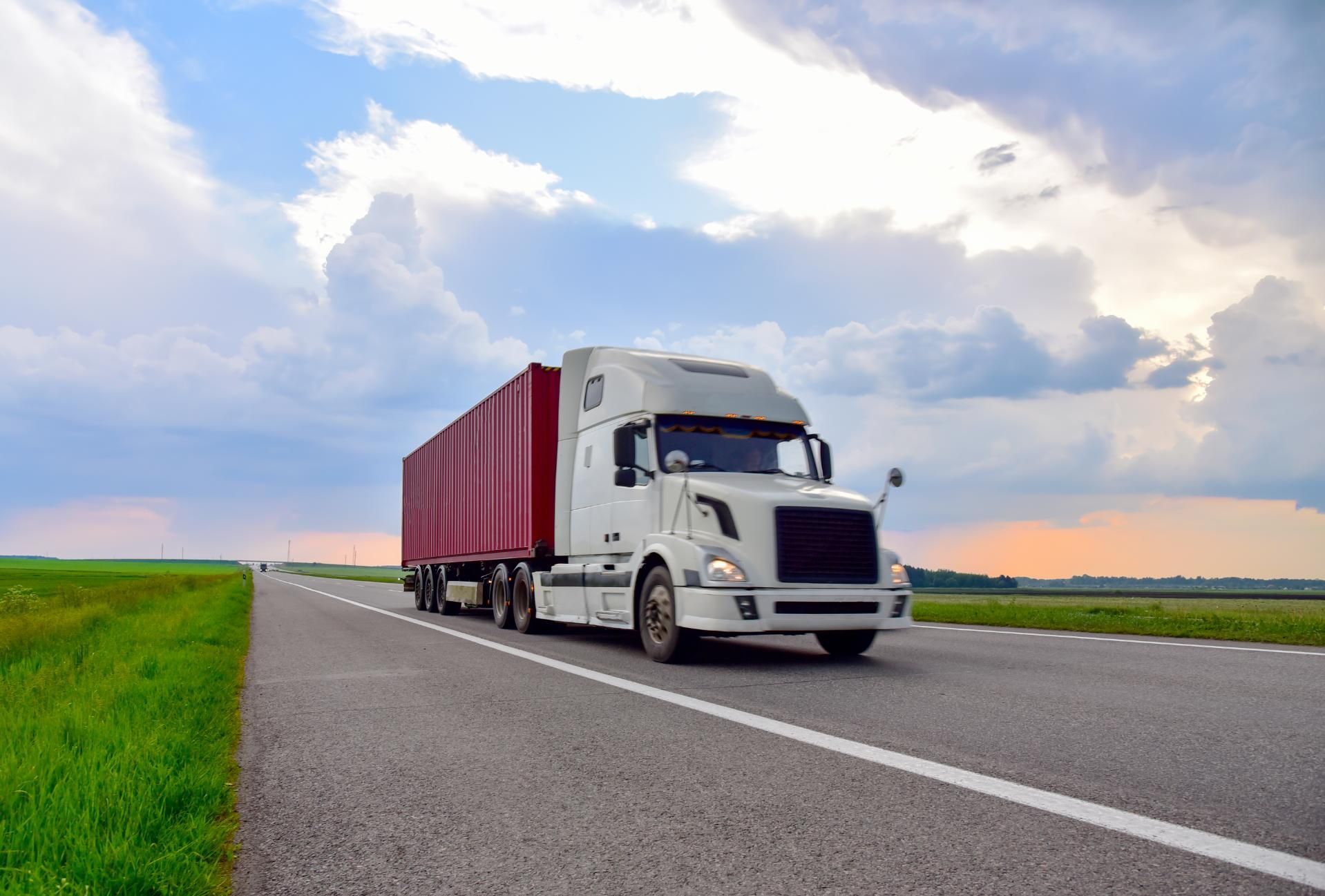 White semi-truck with red trailer driving on a highway, green fields on either side, cloudy sky above.