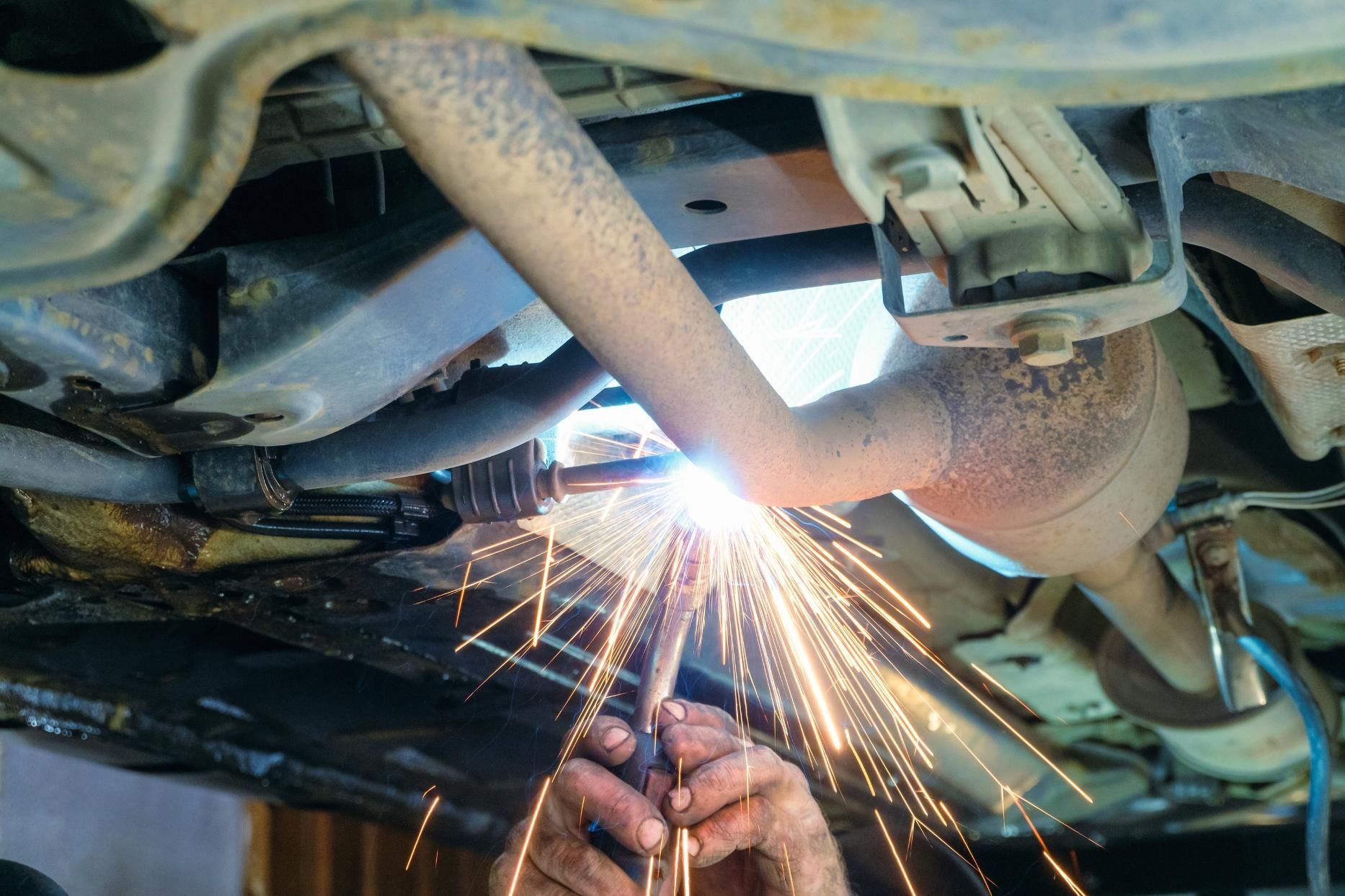 A mechanic welds a car's exhaust pipe, generating sparks under the vehicle.