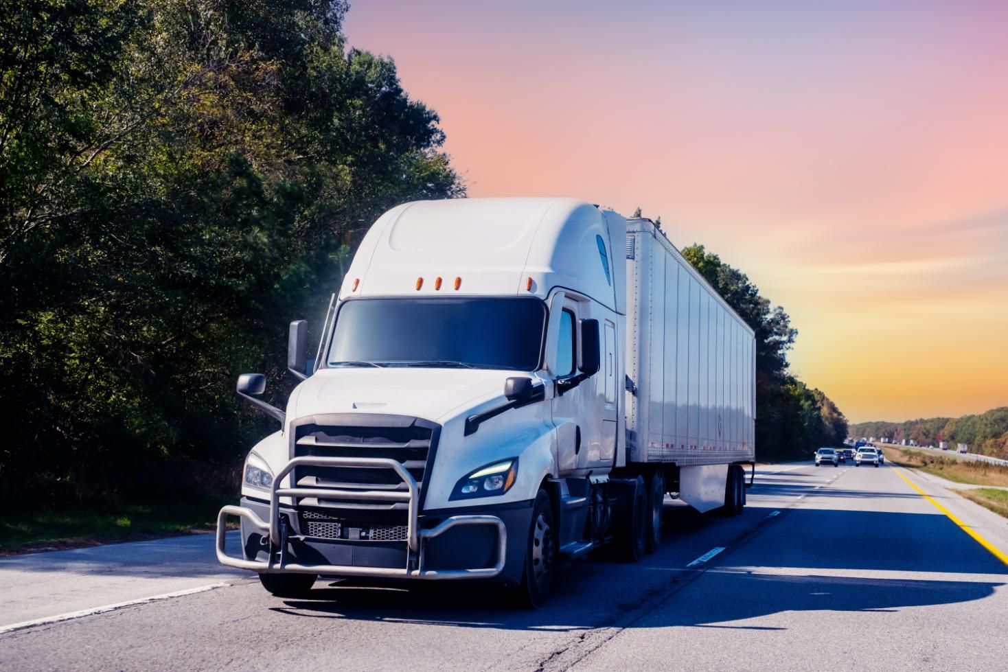 White semi-truck driving on a highway with green trees and a sunset.