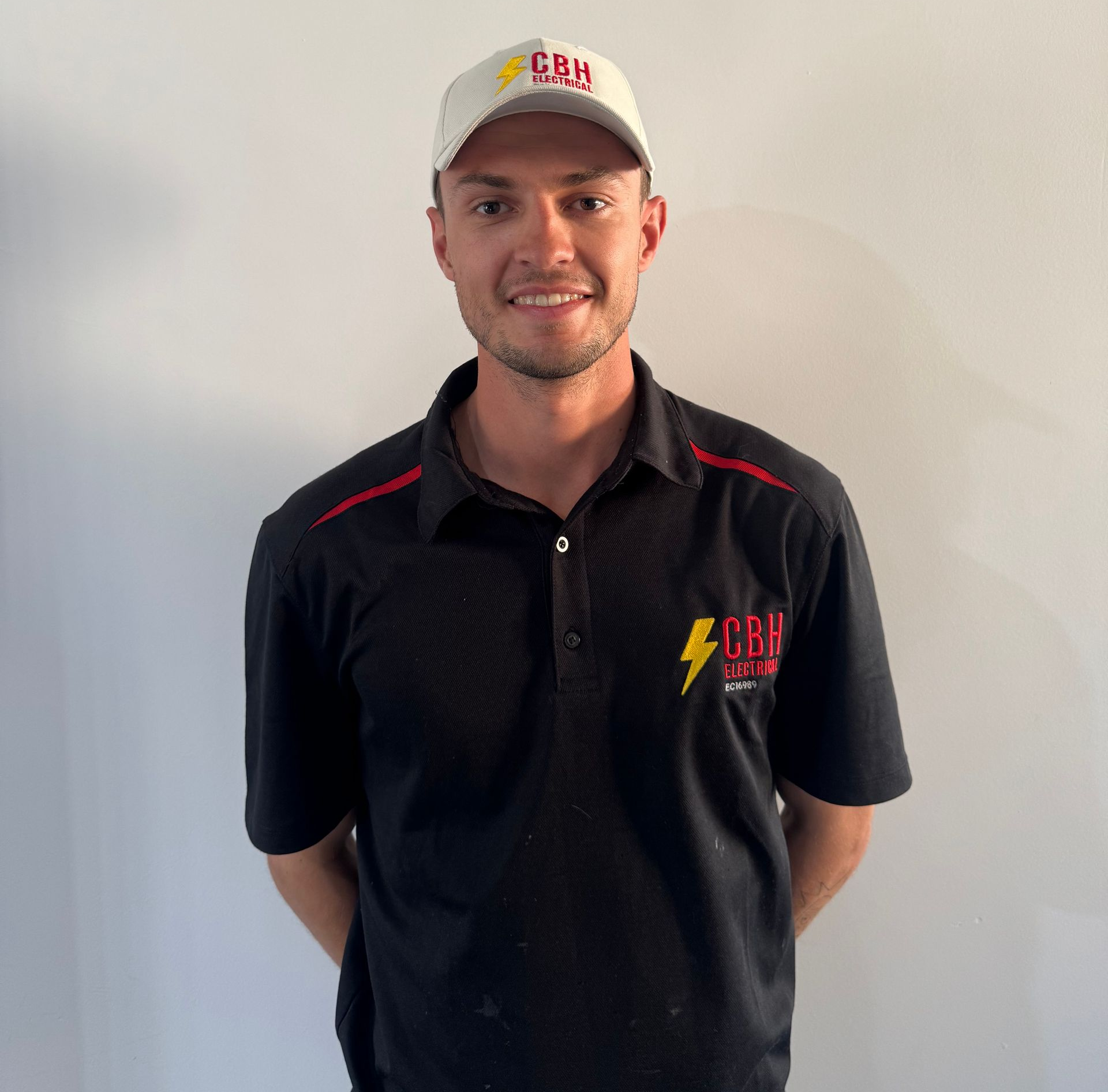 Man in black polo shirt and cap smiling, arms behind back, in front of a white wall.