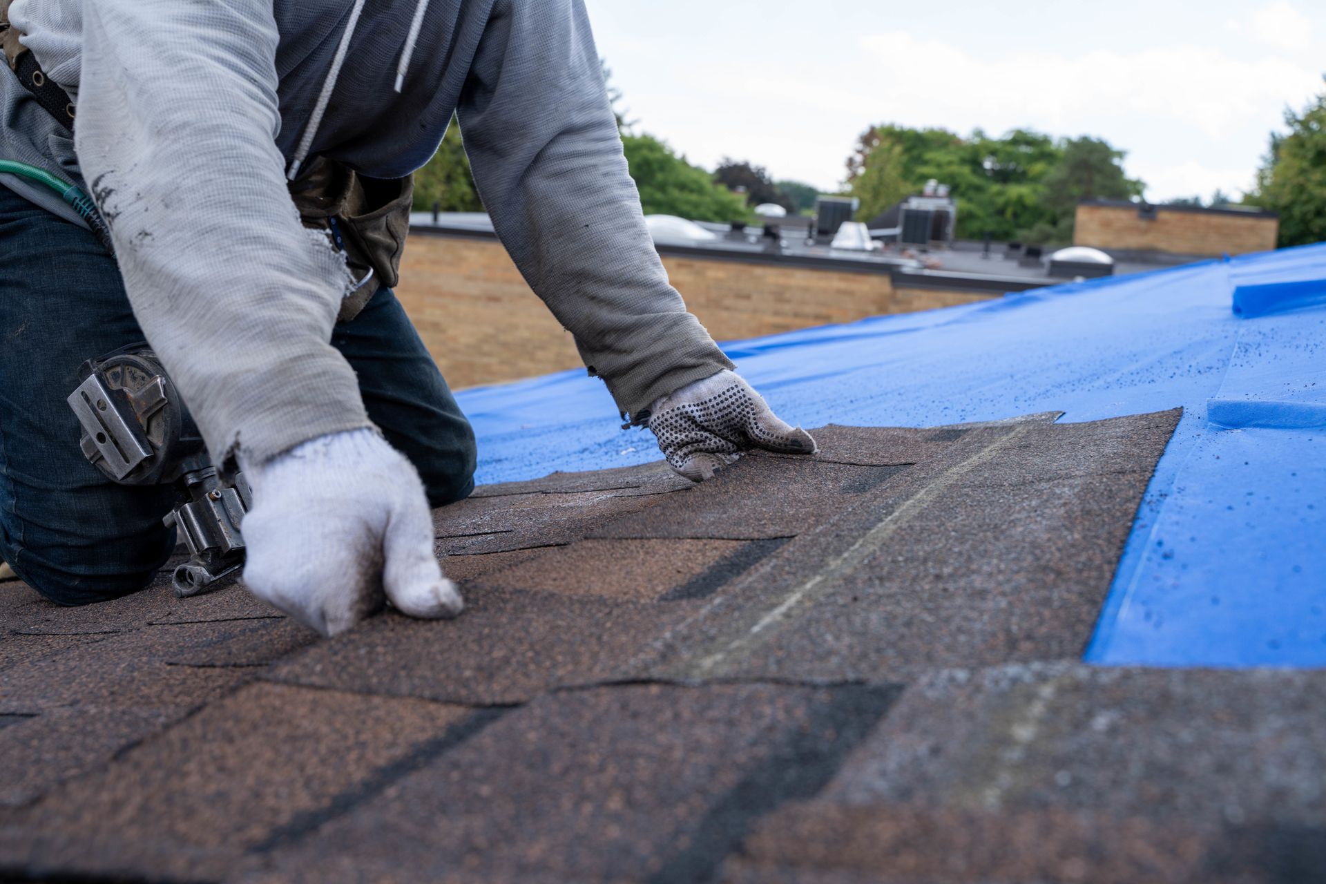 A roofer wearing gloves installs brown asphalt shingles over a blue waterproof underlayment on a roof.