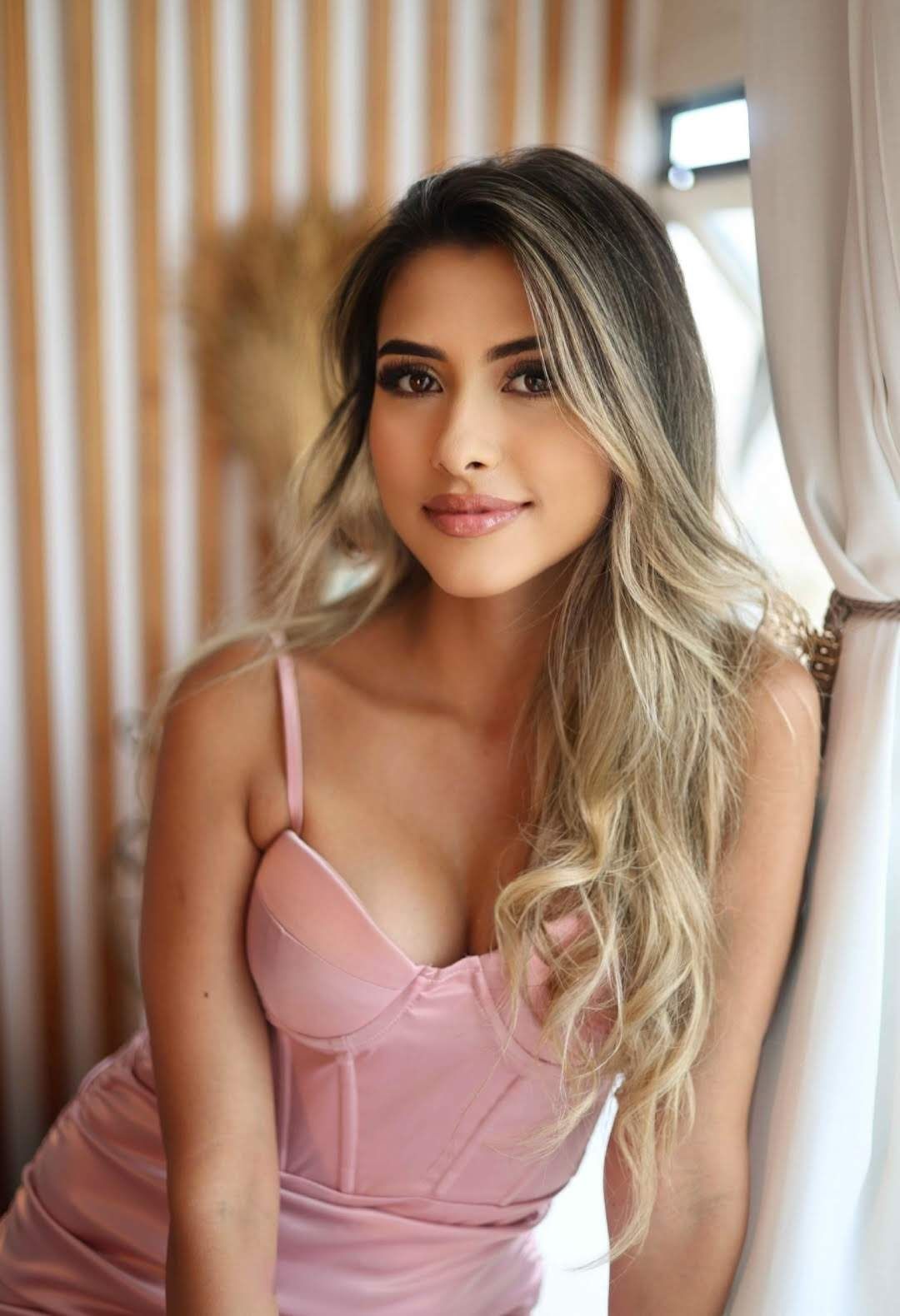 Uma pessoa sorridente, com longos cabelos castanho-claros, vestindo um vestido rosa brilhante estilo corset.