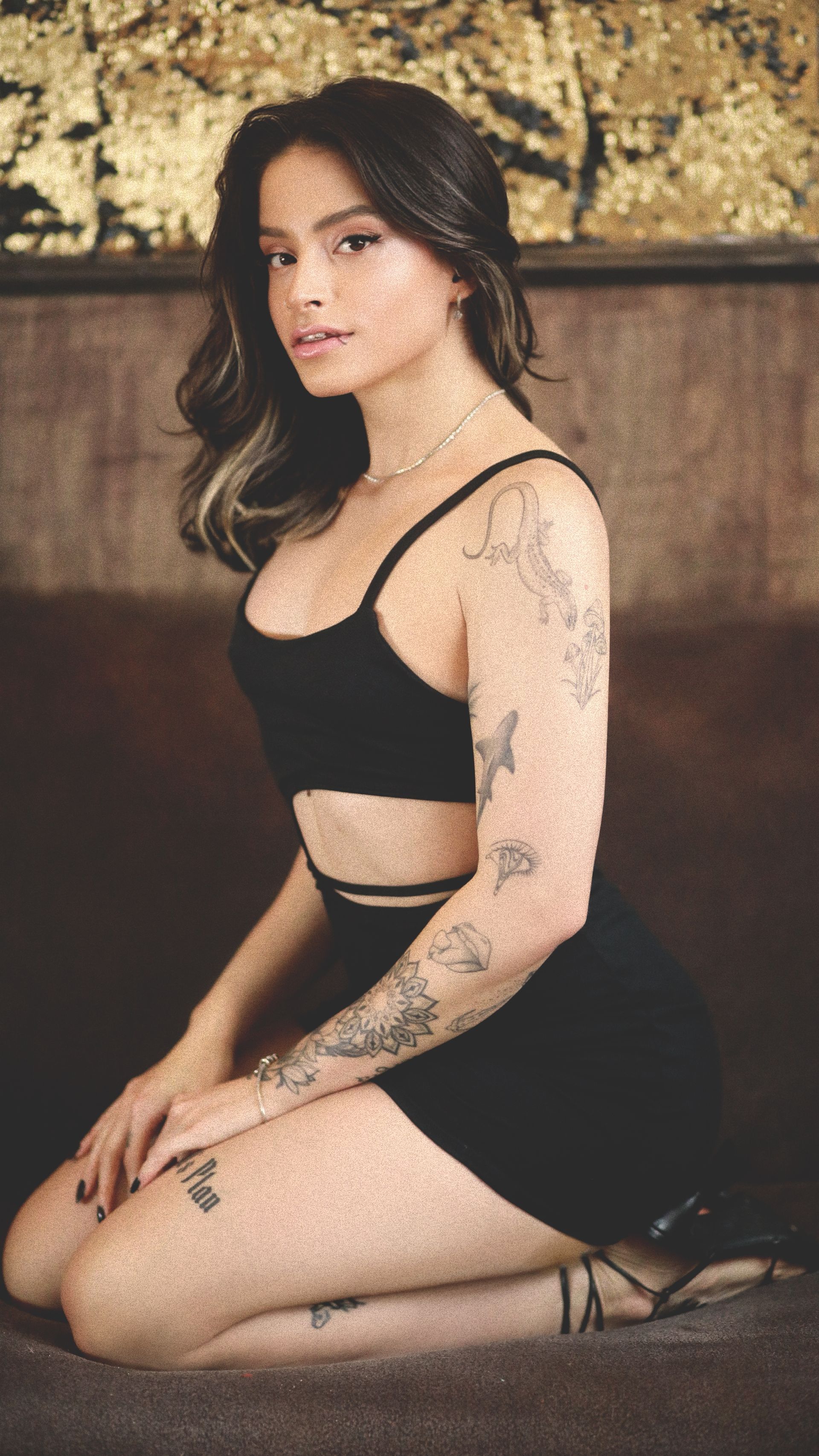 Uma pessoa com tatuagens nos braços e pernas ajoelha-se sobre uma superfície marrom, vestindo um conjunto preto.