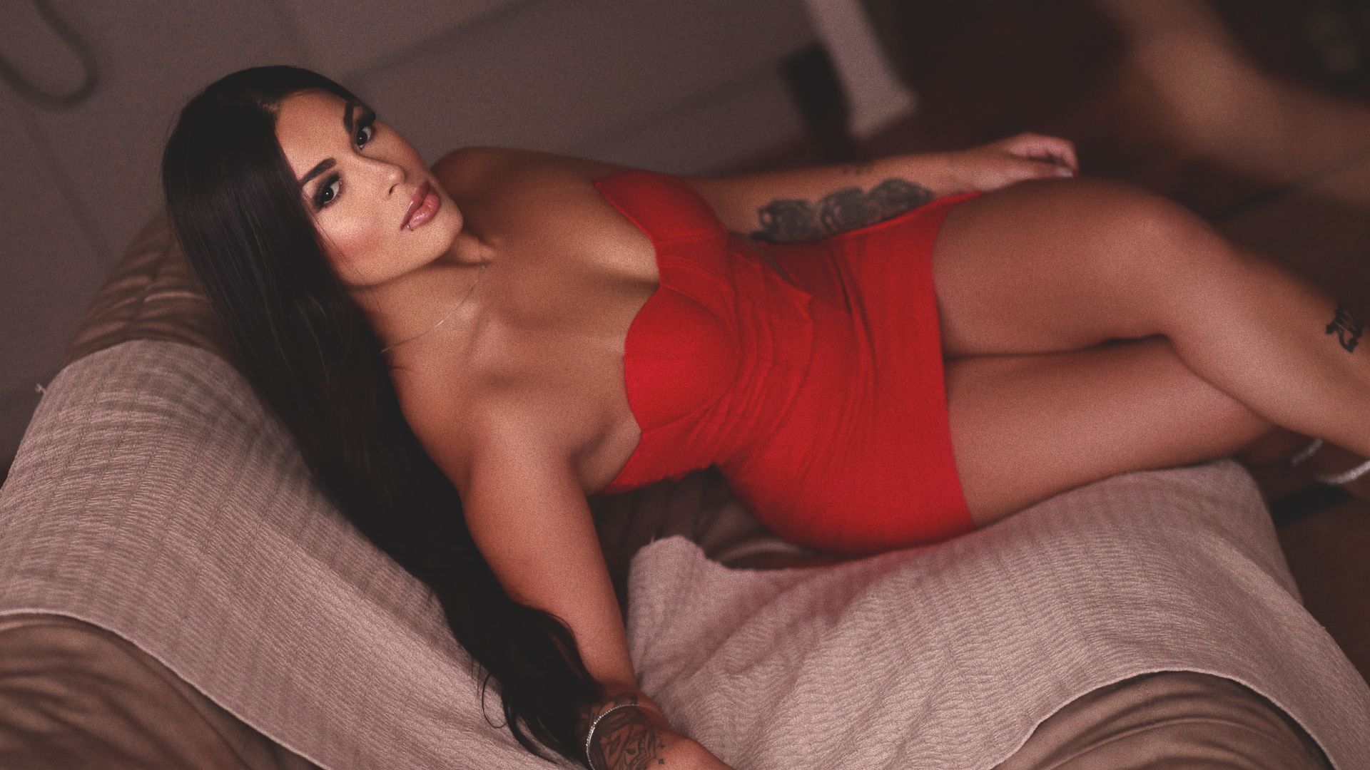 Uma pessoa de cabelos longos e escuros, vestindo um vestido vermelho sem alças, está reclinada em um sofá bege.