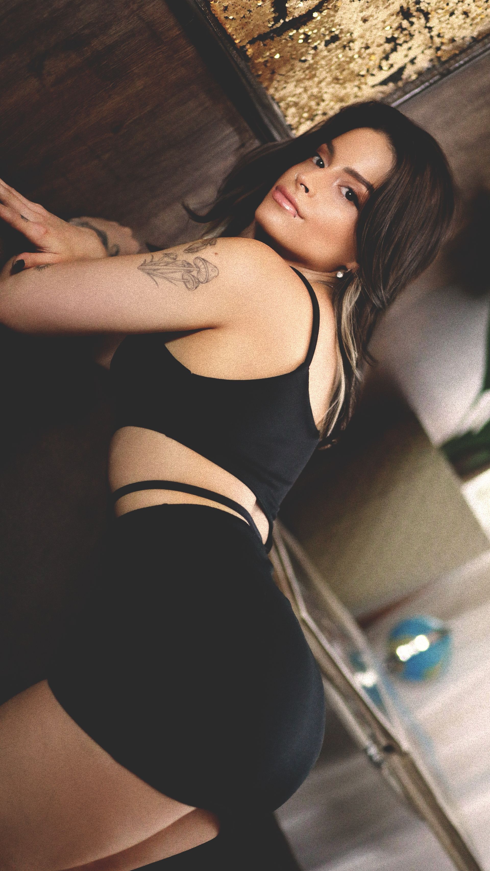 Uma pessoa posando com um top curto preto e shorts, olhando por cima do ombro em direção à câmera em um ambiente interno.