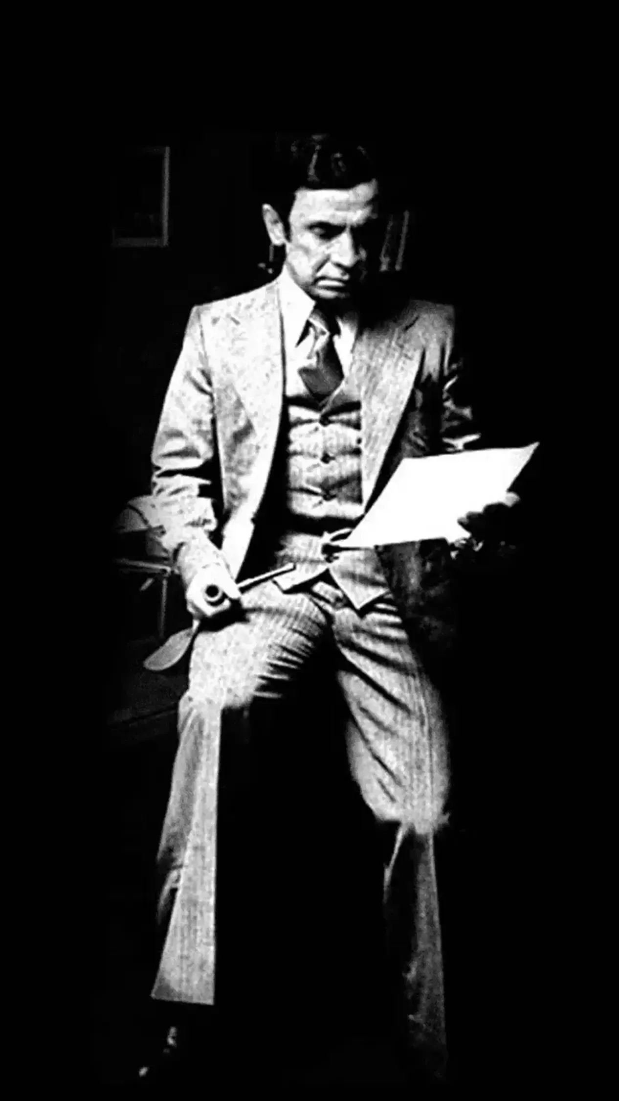 Uma pessoa de terno e colete está sentada em um quarto escuro, olhando para um papel que segura nas mãos.