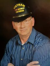 A man wearing a vietnam veteran hat and glasses is sitting with his arms crossed.