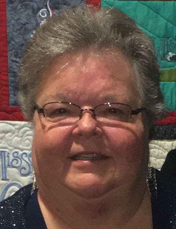 A woman wearing glasses and earrings is smiling in front of a quilt.