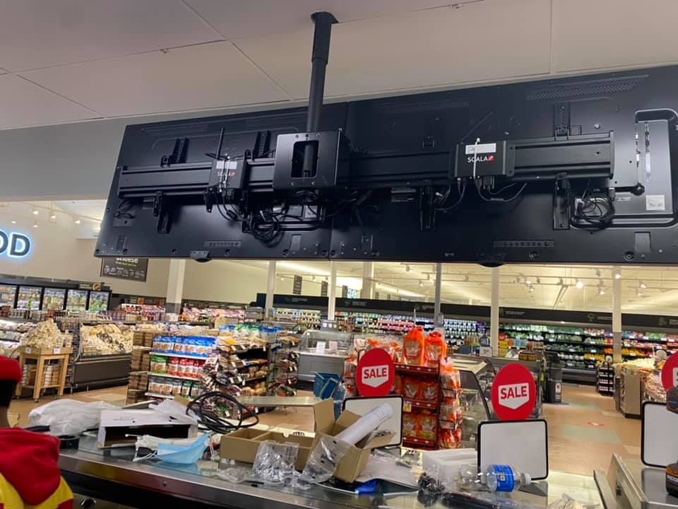 A large flat screen tv is hanging from the ceiling of a grocery store.