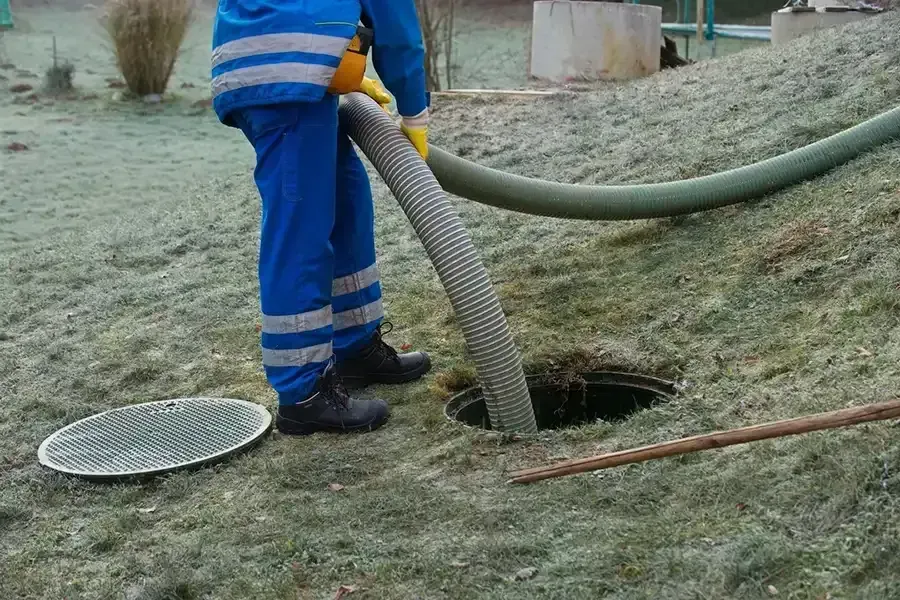 Worker in blue uniform uses a hose to vacuum a hole in the ground outdoors.